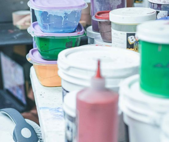 Stacked plastic containers filled with colourful printing inks and various paint buckets on a cluttered workstation, capturing a creative studio environment.