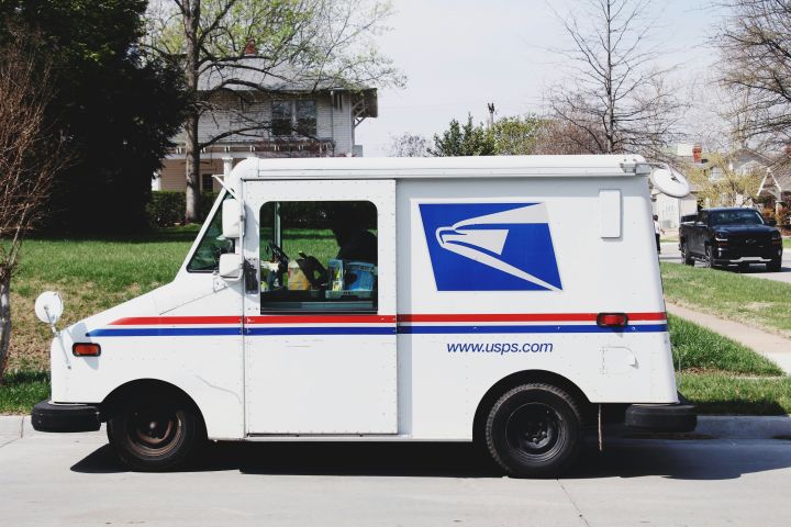 USPS mail delivery truck parked on a residential street, featuring the postal service logo and red and blue stripes, with houses and trees in the background.