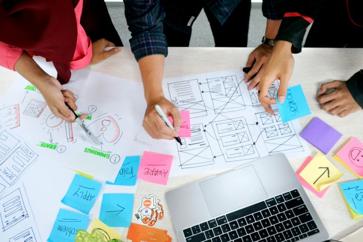 Team collaborating on a design project, sketching wireframes and diagrams on paper, surrounded by colourful sticky notes and a laptop on a desk.