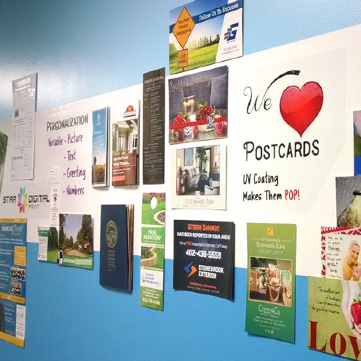 Wall display featuring a variety of printed postcards, brochures, and advertisements with a sign reading 'We ❤️ Postcards' and text promoting UV coating for vibrant designs, set against a light blue background.