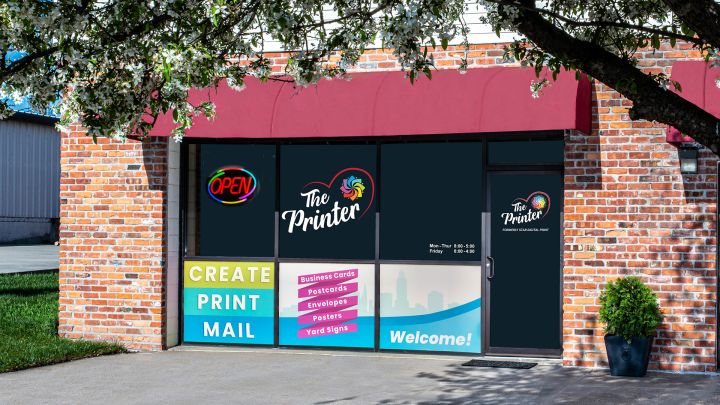 Storefront of The Printer, featuring a red awning, brick exterior, and large windows with colourful signage advertising services like 'Create, Print, Mail' and business cards, postcards, and posters, with a neon 'Open' sign and a welcoming entrance.